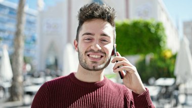 Genç İspanyol adam gülümsüyor. Kafe terasında akıllı telefondan konuşuyor.