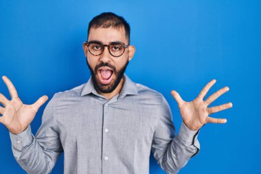Middle east man with beard standing over blue background celebrating crazy and amazed for success with arms raised and open eyes screaming excited. winner concept 