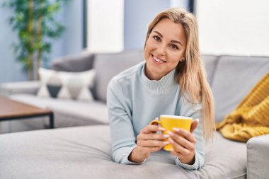 Genç sarışın kadın evdeki kanepede kahve içiyor.