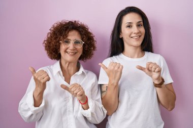 Hispanic mother and daughter wearing casual white t shirt over pink background pointing to the back behind with hand and thumbs up, smiling confident 