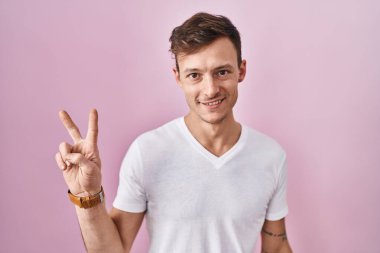 Pembe arka planda duran beyaz adam iki numarayı gösterip işaret ederken kendinden emin ve mutlu bir şekilde gülümsüyor.. 