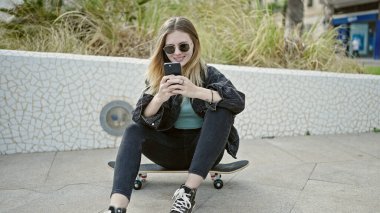 Parkta paten kayarken akıllı telefon kullanan genç sarışın kadın.