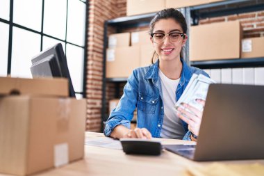 Young beautiful hispanic woman ecommerce business worker holding dollars using calculator at office
