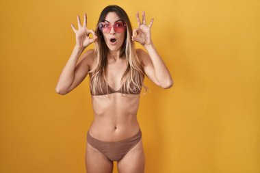 Sarı arka planda bikini giyen genç İspanyol kadın parmaklarıyla onay sembolü yaparken şaşırmış ve şok olmuş görünüyor. çılgın ifade 