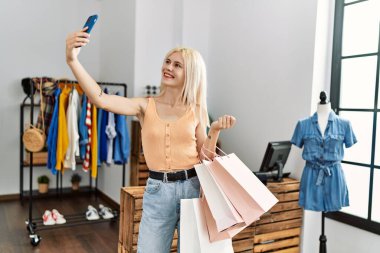 Genç, sarışın, güler yüzlü müşteri elbise mağazasında akıllı telefondan özçekim yapıyor.