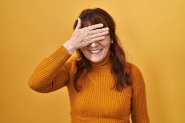 Orta yaşlı İspanyol kadın sarı arka planda duruyor. Gülümsüyor ve gülümsüyor. Gözlerini şaşkınlıkla kapatıyor. kör kavram. 