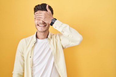 Young hispanic man standing over yellow background smiling and laughing with hand on face covering eyes for surprise. blind concept. 