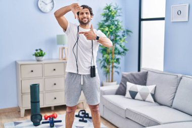 Evde spor kıyafetleri giyen yakışıklı Latin adam gülümsüyor ve mutlu yüzlü eller ve parmaklarla çerçeve yapıyor. yaratıcılık ve fotoğrafçılık kavramı. 