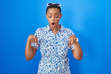 African american woman standing over blue background pointing down with fingers showing advertisement, surprised face and open mouth 