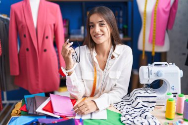 Young beautiful hispanic woman tailor smiling confident sitting on table at clothing factory