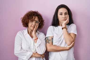 İspanyol anne ve kızı pembe arka plan üzerine gündelik beyaz tişört giyip kollarını kavuşturarak depresyondan sıkılmış ve yorgun görünüyorlar.. 
