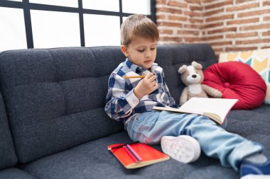 Evdeki kanepede oturan beyaz çocuk defterine resim çiziyor.