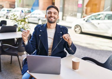 Laptop kullanan, kahve içen, kahve dükkanının terasında oturan Latin kökenli genç adam.