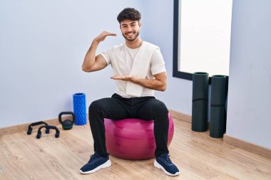 Sakallı İspanyol bir adam yoga odasında pilatese oturmuş büyük ve büyük boy işareti gösteren el hareketleriyle ölçü sembolü gösteriyor. Kameraya bakıp gülümsüyor. ölçüm kavramı. 