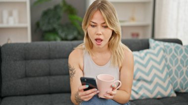 Genç sarışın kadın akıllı telefon kullanıyor. Evde sürpriz bir ifadeyle kahve içiyor.