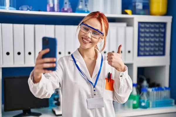 科学者研究所で働く若い白人女性がスマートフォンの笑顔でビデオ通話を行います幸せと正 親指アップは優れたと承認サインを行います — ストック写真