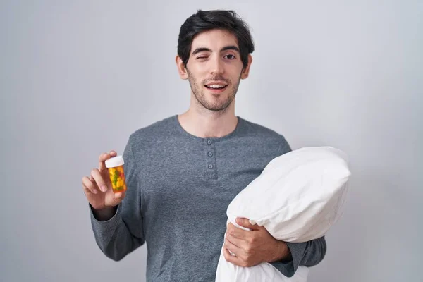 Young Hispanic Man Wearing Pyjama Hugging Pillow Holding Pills Winking — Stock Photo, Image