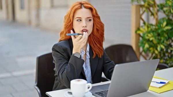 コーヒーショップのテラスでスマホから音声メッセージを送るノートパソコンを利用した若い赤毛の女性ビジネスワーカー — ストック写真
