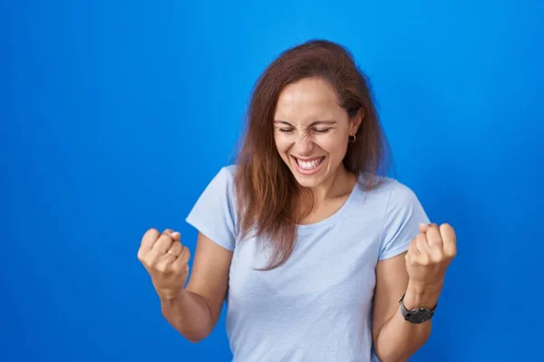 布鲁内特的女人站在蓝色的背景上 非常高兴而兴奋地做着获胜的手势 举起双臂 微笑着 尖叫着要成功 庆祝概念 — 图库照片