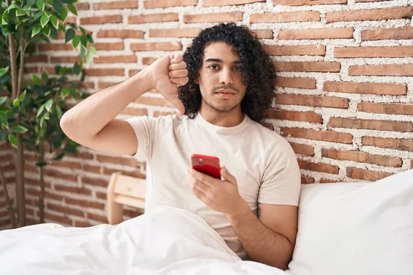 Ισπανόφωνος Άνθρωπος Σγουρά Μαλλιά Χρησιμοποιώντας Smartphone Κάθεται Στο Κρεβάτι Θυμωμένος — Φωτογραφία Αρχείου