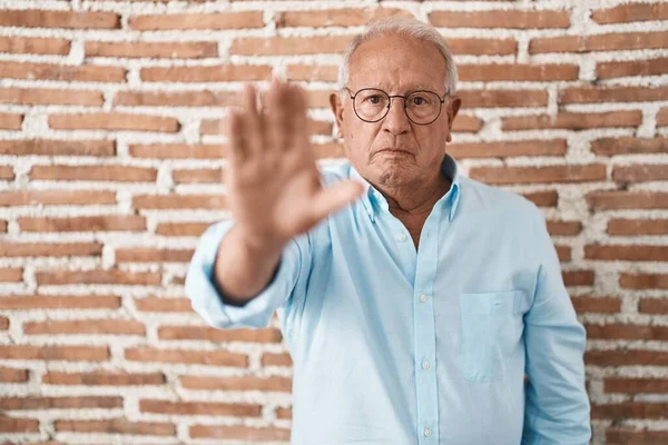 Senior man with grey hair standing over bricks wall doing stop sing with palm of the hand. warning expression with negative and serious gesture on the face.