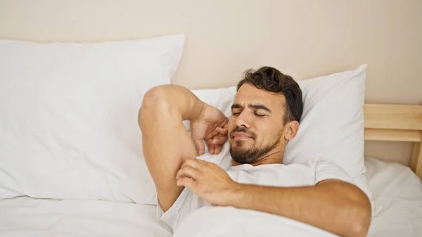 Young hispanic man lying on bed scratching arm at bedroom