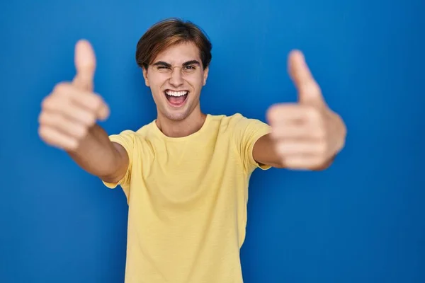 年轻男子站在蓝色的背景上 赞成用手做积极的手势 微笑着竖起大拇指 为成功感到高兴 优胜手势 — 图库照片
