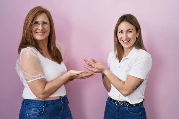 西班牙裔母亲和女儿身穿宽松的白色T恤 背景粉色 手拉手张开手掌 展示复制空间 呈现广告笑容 兴奋极了 — 图库照片