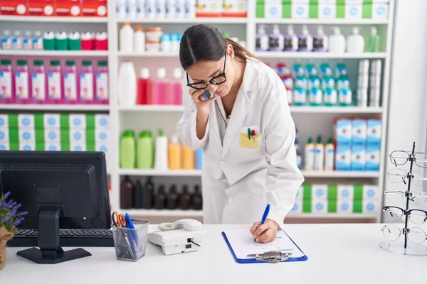 Young Beautiful Hispanic Woman Pharmacist Talking Smartphone Writing Document Pharmacy — Stok fotoğraf
