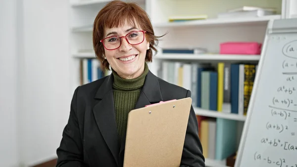 stock image Mature hispanic woman teacher standing by white board with clipboard at library university