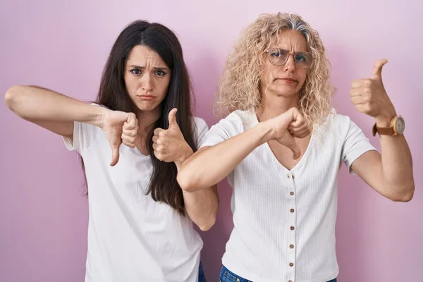 Mother and daughter standing together over pink background doing thumbs up and down, disagreement and agreement expression. crazy conflict