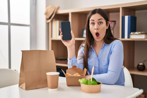 年轻的黑发女人在家里吃东西时 带着食物回家 显示出智能手机屏幕的恐惧和惊讶 张大了嘴表示惊奇和怀疑 — 图库照片
