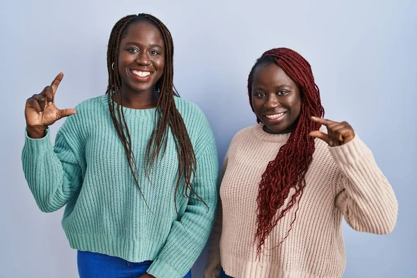 青い背景の上に立つ2人のアフリカ人女性は笑顔で 指を見てカメラで小さなサイズのサインをする手で自信を持ってジェスチャー 概念を測定し — ストック写真