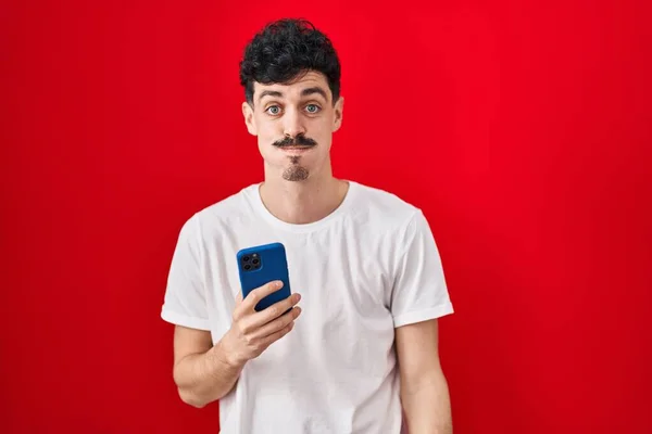 Uomo Ispanico Utilizzando Smartphone Sfondo Rosso Sbuffare Guance Con Faccia — Foto Stock