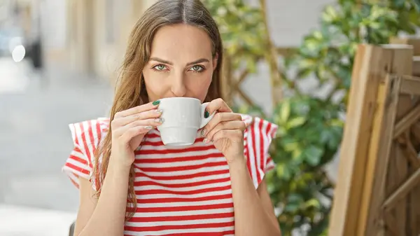 Young blonde woman drinking cup of coffee sitting on table at coffee shop terrace