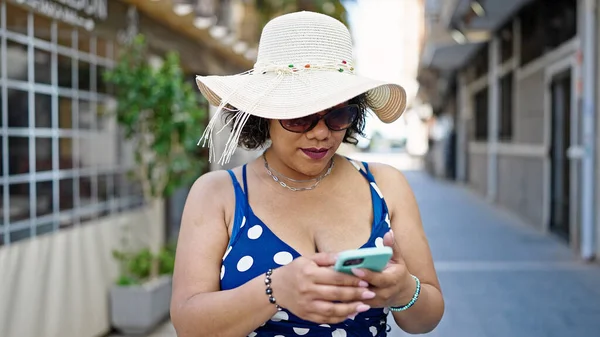 Sokağın ortasında akıllı telefon kullanarak yaz şapkası ve güneş gözlüğü takan genç, güzel Latin kadın turist.