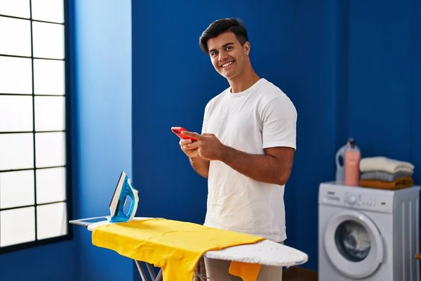 Ung Latinamerikan Man Med Smartphone Strykning Kläder Tvättstuga — Stockfoto