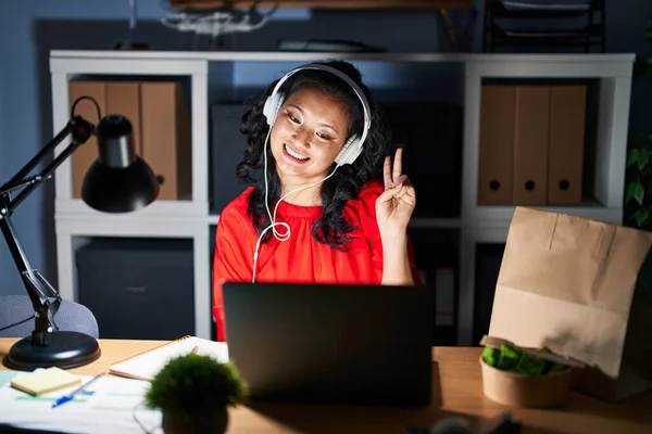 夜はノートパソコンを持ってオフィスで働く若いアジア人女性が勝利サインをしてカメラに顔をウィンクして微笑む — ストック写真