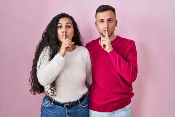 Young Hispanic Couple Standing Pink Background Asking Quiet Finger Lips — ストック写真