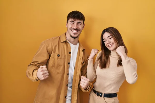 黄色の背景に立っている若いヒスパニック系のカップルは非常に満足し 腕を上げて勝者のジェスチャーを行う興奮し 笑顔と成功のために叫んで お祝いのコンセプト — ストック写真