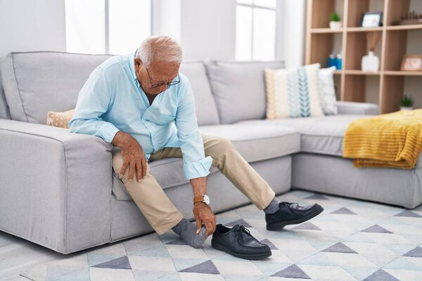 Senior grey-haired man suffering for feet pain sitting on sofa at home