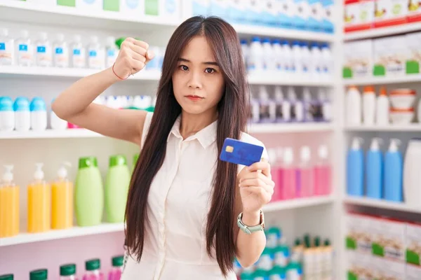 Young chinese woman shopping at pharmacy drugstore holding credit card annoyed and frustrated shouting with anger, yelling crazy with anger and hand raised