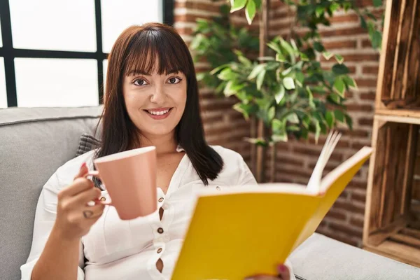 Young Beautiful Hispanic Woman Reading Book Drinking Coffee Sitting Sofa —  Fotos de Stock
