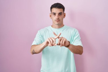Yakışıklı İspanyol adam pembe arka planda duruyor. Reddedilme yüz ifadesi. Eksi işareti yapıyor. 