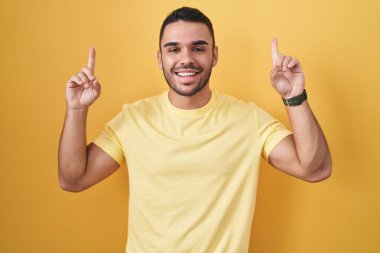 Young hispanic man standing over yellow background smiling amazed and surprised and pointing up with fingers and raised arms. 