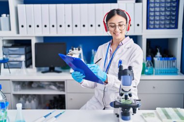 Young hispanic woman scientist listening to music writing on document at laboratory