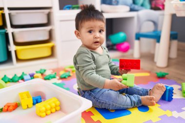 Adorable hispanic boy playing with construction blocks sitting on floor at kindergarten
