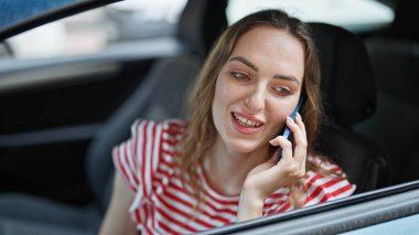 Genç sarışın kadın akıllı telefondan konuşuyor. Sokakta arabada oturuyor.