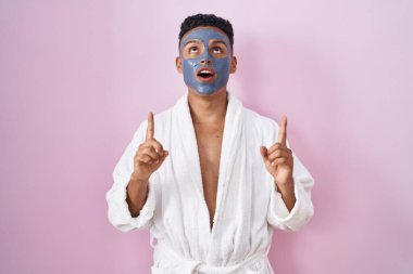 Güzellik maskesi ve banyo cübbesi takan İspanyol genç adam hayret ve şaşkınlık içinde yukarı baktı ve parmaklarıyla işaret etti ve kollarını kaldırdı.. 