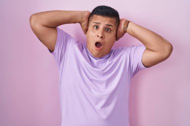 Young hispanic man standing over pink background crazy and scared with hands on head, afraid and surprised of shock with open mouth 